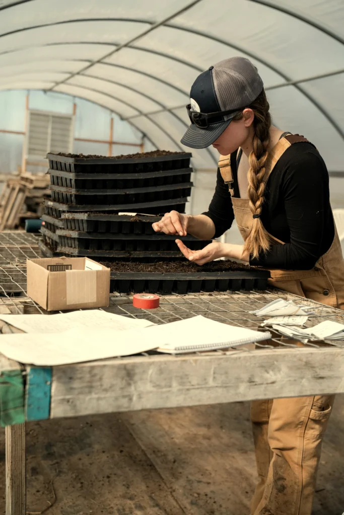 Grower in overalls planting seedlings into black nursery trays inside a greenhouse, Santa Fe NM