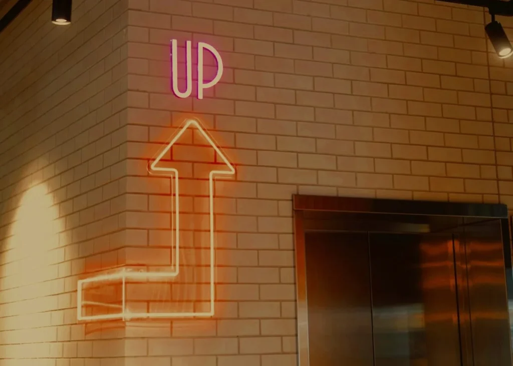 A neon 'UP' sign with an upward arrow beside an elevator door, the momentum Kingster Local's Business Accelerator creates.